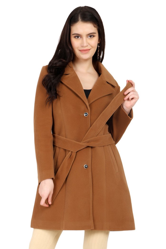 A woman is giving a standing pose, wearing Trufit’s Long Dk. Camel Velvet coat with a lapel collar, button closure, belt, side pockets, and skin bell bottom and holding the coat’s belt.